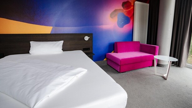 Ein Hotelzimmer mit einem großen Bett, einem rosafarbenen Sofa, einem kleinen weißen Tisch und einem farbenfrohen abstrakten Wandbild.