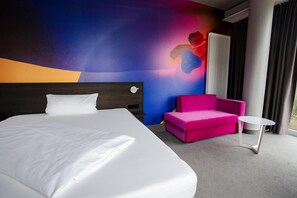 Une chambre d’hôtel avec un grand lit, un canapé rose, une petite table blanche et une fresque murale abstraite et colorée.