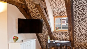 A cozy room with a flat-screen TV mounted on the wall, a small table with chairs, a white cabinet, and a window with a view of the outside. - Hotel David (Regensburg)