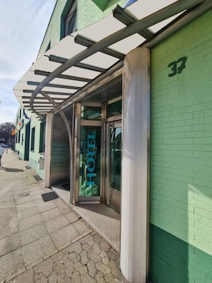 Entrada de hotel con puerta de vidrio, toldo de metal y pared de ladrillo verde.