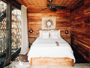 A wooden cabin bedroom with a bed, a ceiling fan, and a window with curtains. - Nomade Holbox (Isla Holbox)