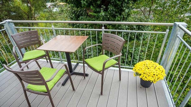 Une terrasse en bois avec une table et deux chaises, une rambarde en métal et un pot de fleurs contenant des fleurs jaunes.