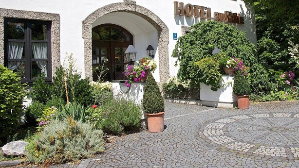 Ein Hotel mit Eingangsbereich, Kopfsteinpflasterauffahrt, gewölbten Durchgängen und Kübelpflanzen.