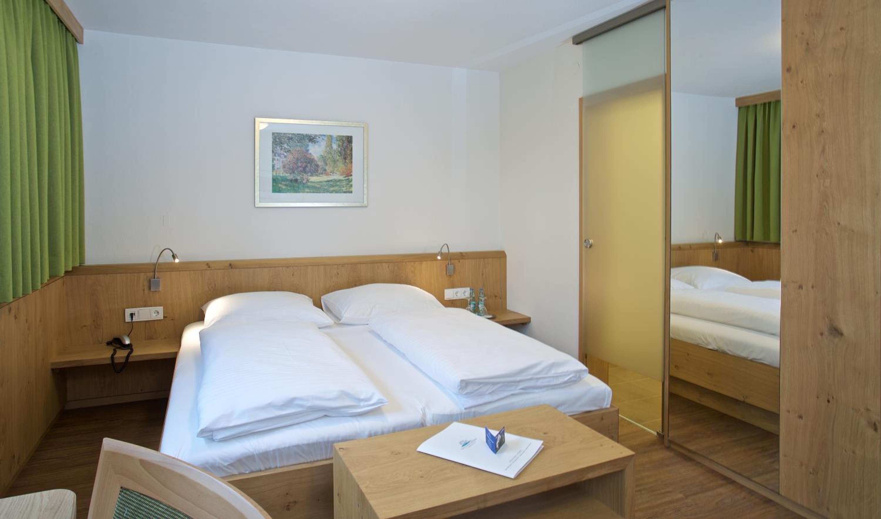 Een hotelkamer met een houten hoofdeinde, een bed met witte lakens, een nachtkastje met een lamp, een klein bureau en een groen gordijn.