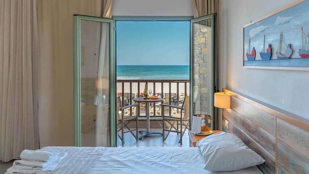 Een hotelkamer met een bed, een tafel met stoelen, een schilderij en uitzicht op zee.