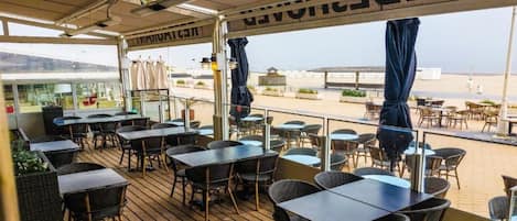 Et udendørs spiseområde med borde og stole, udsigt til stranden og en overdækket gang.