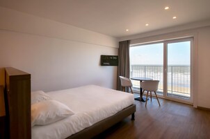 Een hotelkamer met een bed, een bureau en een stoel. Er is een groot raam met uitzicht op zee.