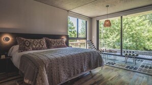 A modern bedroom with a large bed, a nightstand, a window with a view of trees, and a balcony with chairs. - Be House Suites & Spa by Nobile (Punta del Este)