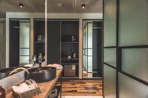 A modern kitchen with a wooden countertop, a large black pan, and a coffee machine. There are cabinets with glass doors and a view of a hallway with a green screen. - Be House Suites & Spa by Nobile (Punta del Este)