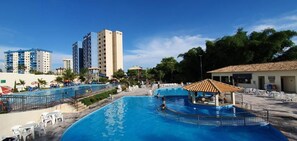 A swimming pool with a sunshade, surrounded by lounge chairs and umbrellas, with tall buildings in the background. - Suite Para ATE 4 Pessoas EM Golden Dolphin Grande Hotel Piscina 24h (Caldas Novas)