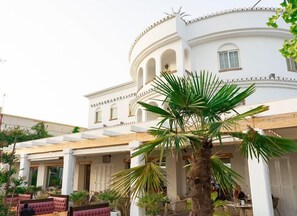 Um edifício branco com fachada curva, colunas e uma área externa coberta com cadeiras e mesas acolchoadas.