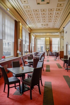Un comedor de hotel con mesas, sillas, ventanas amplias y una alfombra roja.