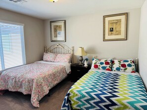 A bedroom with two beds, each with a distinct patterned bedspread. There are two framed pictures on the wall, a bedside table with a lamp, and a window with blinds.