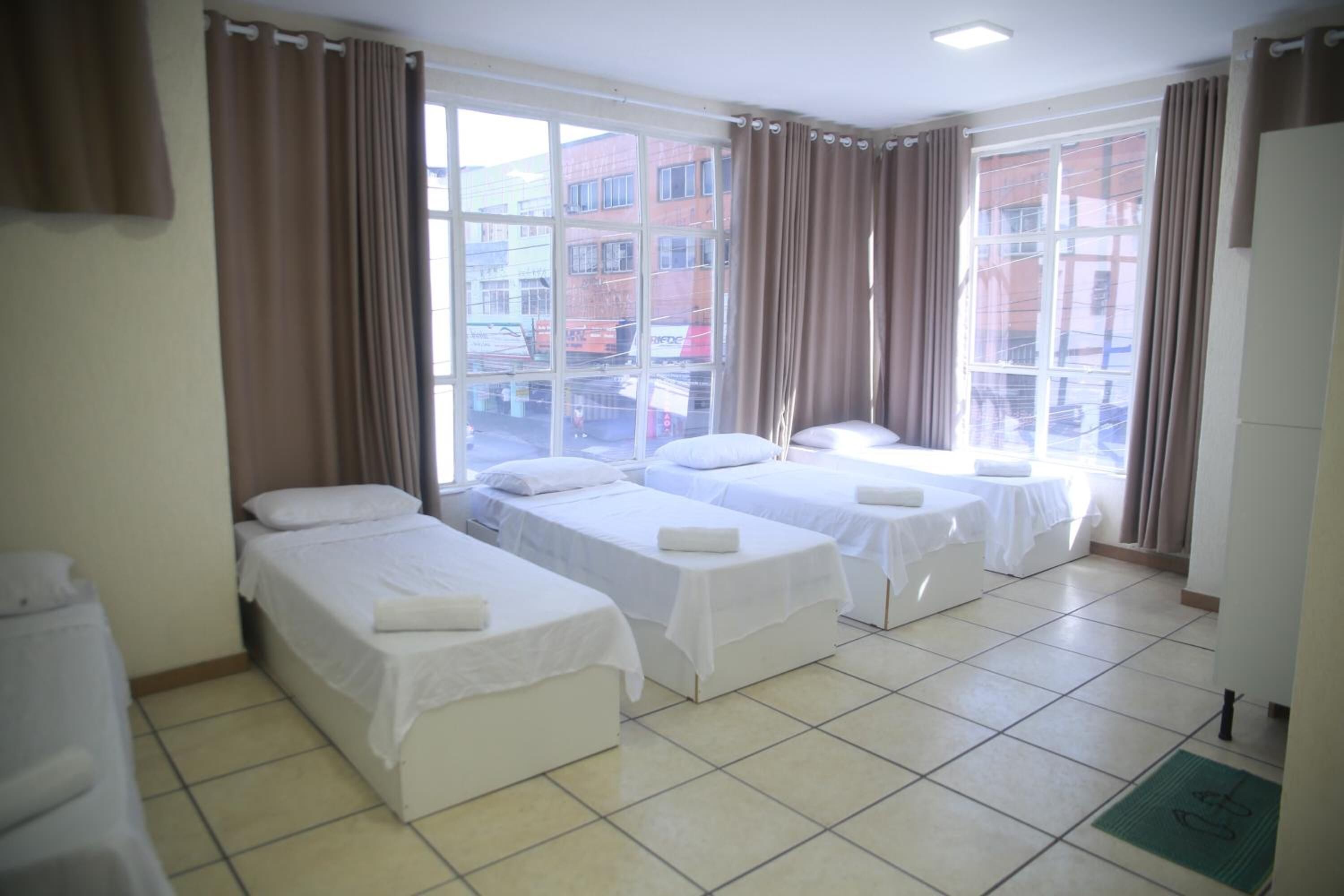 A hotel room with three single beds, large windows with curtains, and a tiled floor.