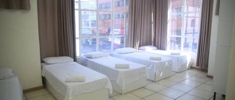 A hotel room with three single beds, large windows with curtains, and a tiled floor.