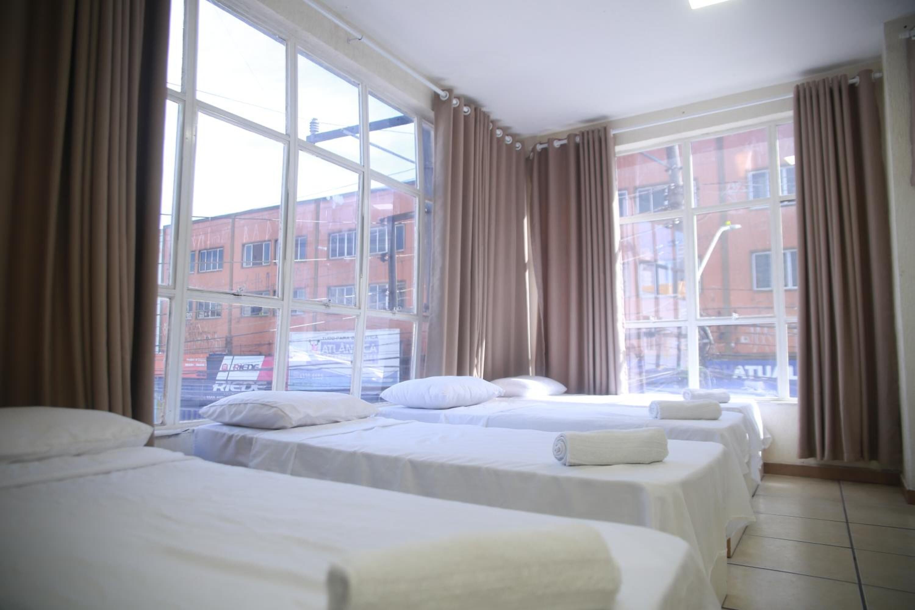 Quarto de hotel com três camas, janelas amplas e cortinas.