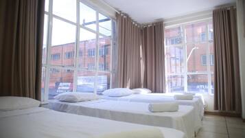 Quarto de hotel com três camas, janelas amplas e cortinas.