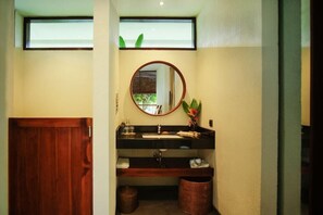 A bathroom with a wooden door, a round mirror, a sink with a black countertop, and a window above the mirror. - Ama-lurra Resort Gili Air (Gili Air)