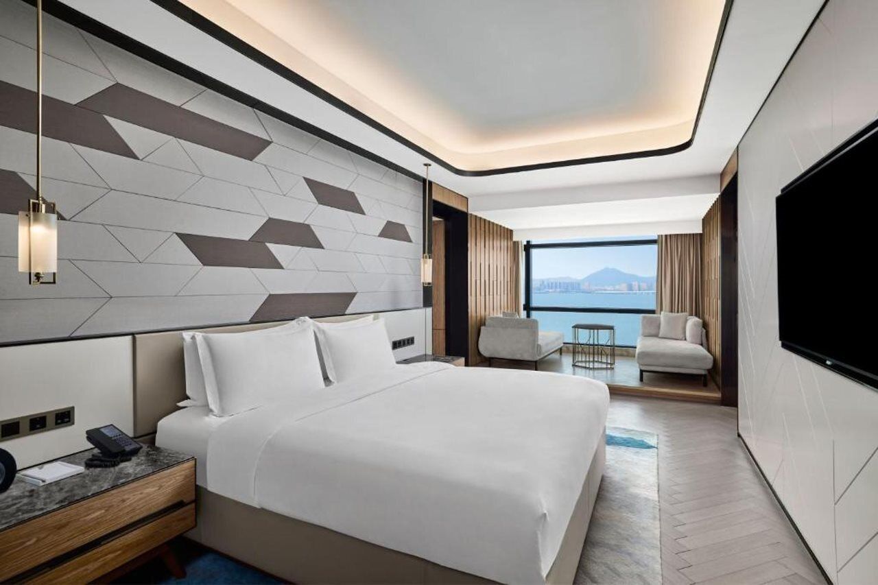 A modern hotel room with a large bed, a desk with a phone, a TV, and a view of the city.