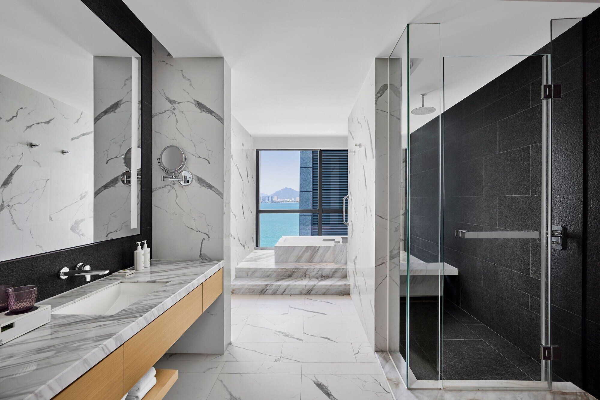 A modern bathroom with a marble vanity, a large mirror, a glass-enclosed shower, and a view of the sea.
