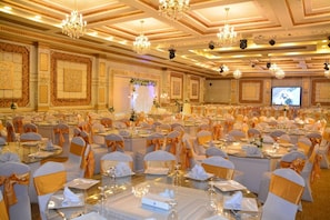 A banquet hall with round tables, chairs with orange sashes, chandeliers, and a large screen. - Rehana Resort (Cairo)