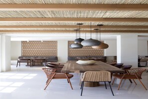 A spacious dining area with a long wooden table, multiple chairs, and hanging pendant lights. - Elessa Hotel (Santorini)