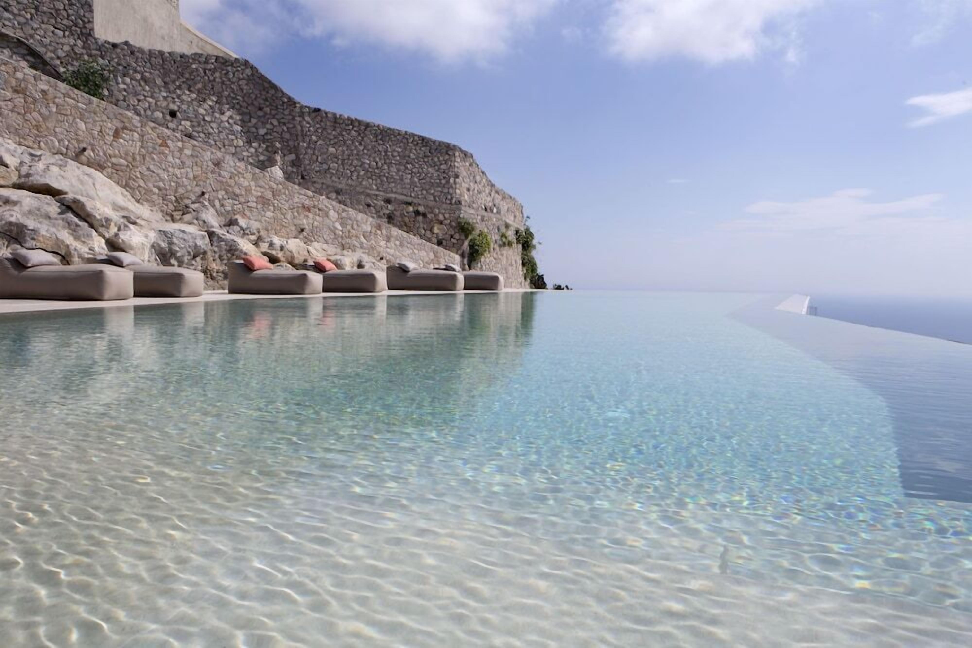 Una piscina infinita cristalina con un muro de piedra y sillones de descanso.