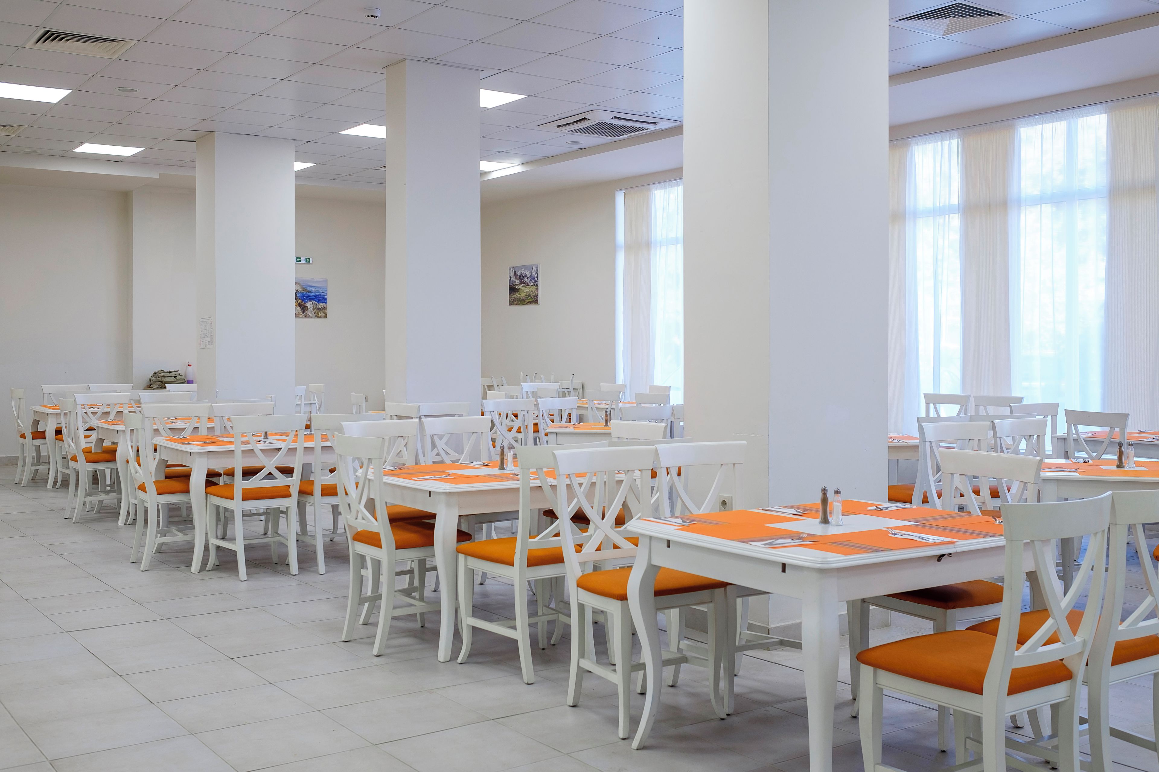 A dining area with white tables and chairs, arranged in rows, with large windows allowing natural light.