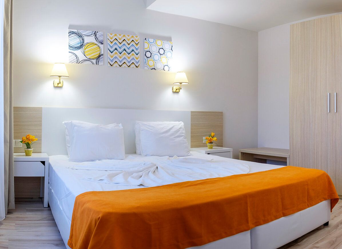 A hotel room with a bed, two bedside tables, a nightstand, a wardrobe, and wall art.