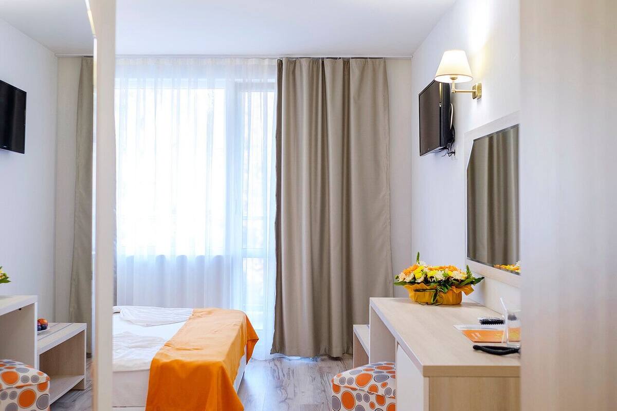 A hotel room with a bed, a desk, a chair, a mirror, a window with curtains, and a small table with a flower vase.
