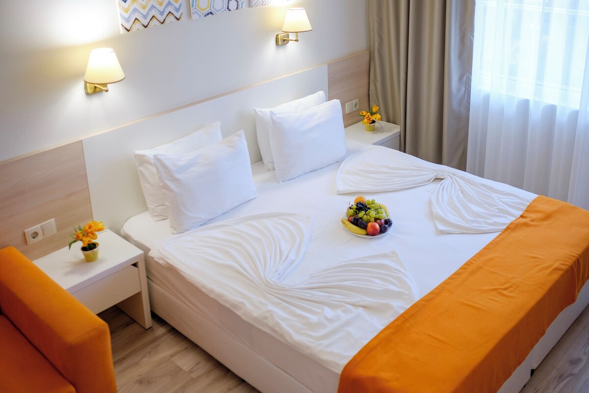 Una habitación de hotel con una cama bien tendida, una mesita de noche con lámpara, un pequeño jarrón con flores y un frutero en la cama.