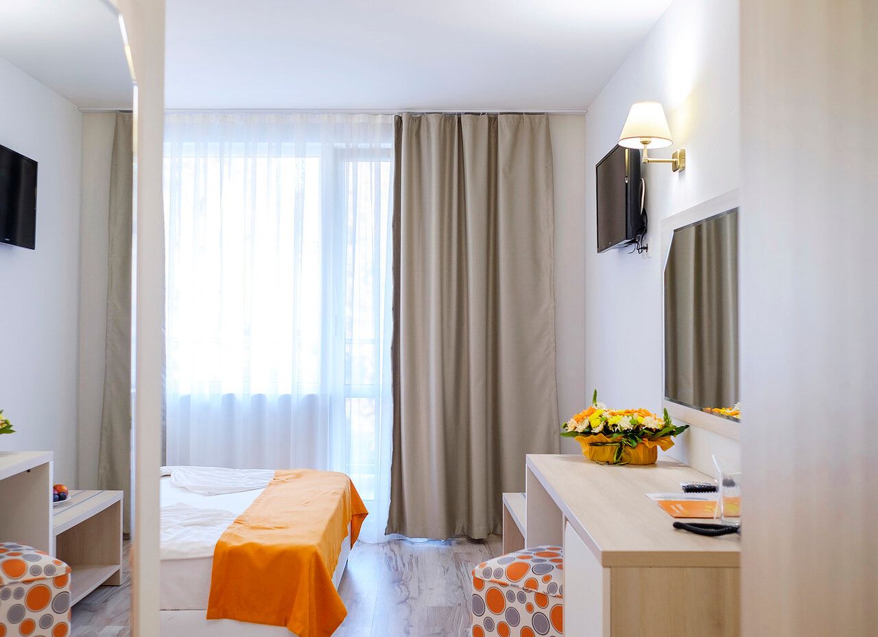 Une chambre d’hôtel comprenant un lit, un bureau, une chaise, un miroir, une fenêtre avec des rideaux et une petite table sur laquelle est posé un vase avec une fleur.