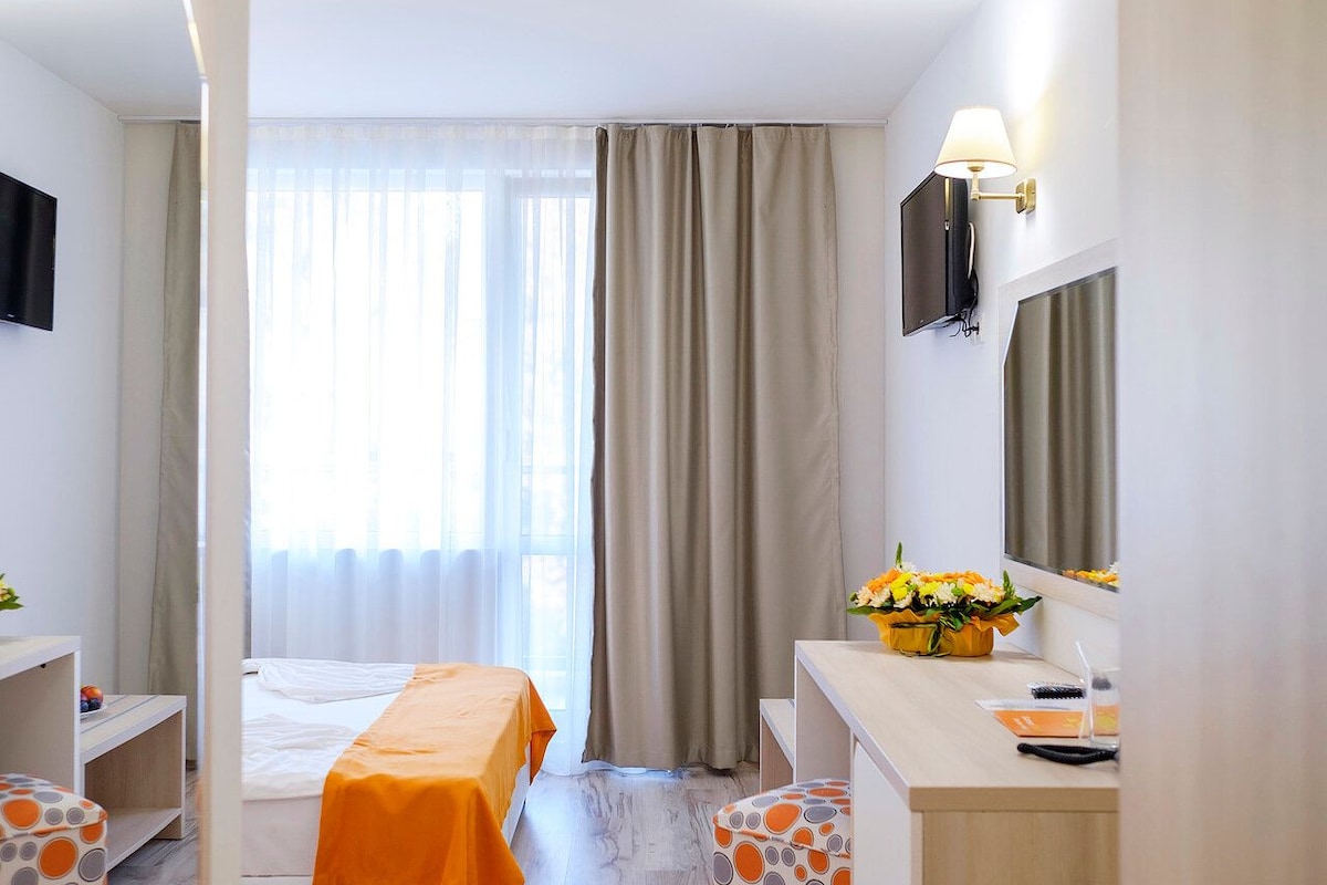 Habitación de hotel con cama, escritorio, silla, espejo, ventana con cortinas y una mesita con un florero.