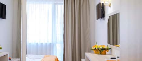 A hotel room with a bed, a desk, a chair, a mirror, a window with curtains, and a small table with a flower vase.