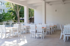 Un área de comedor al aire libre con mesas y sillas blancas, una sección cubierta y vistas a edificios y árboles.