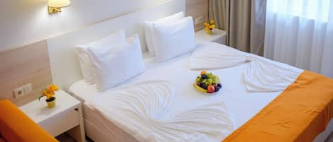 A hotel room with a neatly made bed, a bedside table with a lamp, a small vase with flowers, and a fruit bowl on the bed.
