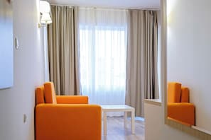 Una habitación con una mesa blanca, dos sillas naranjas, una ventana con cortinas de gasa y una lámpara fijada a la pared.