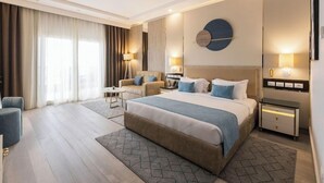 A modern hotel room with a large bed, a sitting area with a sofa and armchairs, a desk, and a wall-mounted round mirror. - Gewan Resort New Alamein (El Alamein)