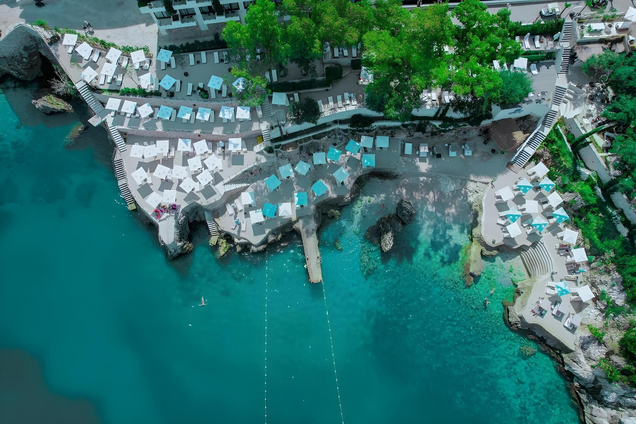 An aerial view of a coastal resort with multiple beachside lounge areas, clear turquoise waters, and rocky formations.