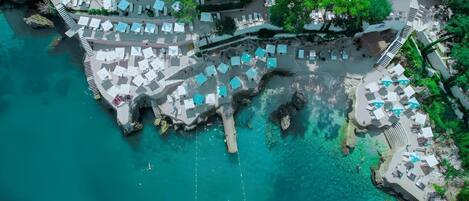 An aerial view of a coastal resort with multiple beachside lounge areas, clear turquoise waters, and rocky formations.