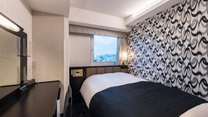 A hotel room with a bed, a desk, a mirror, a window with a city view, and patterned wallpaper. - Apa Hotel (Tokyo)