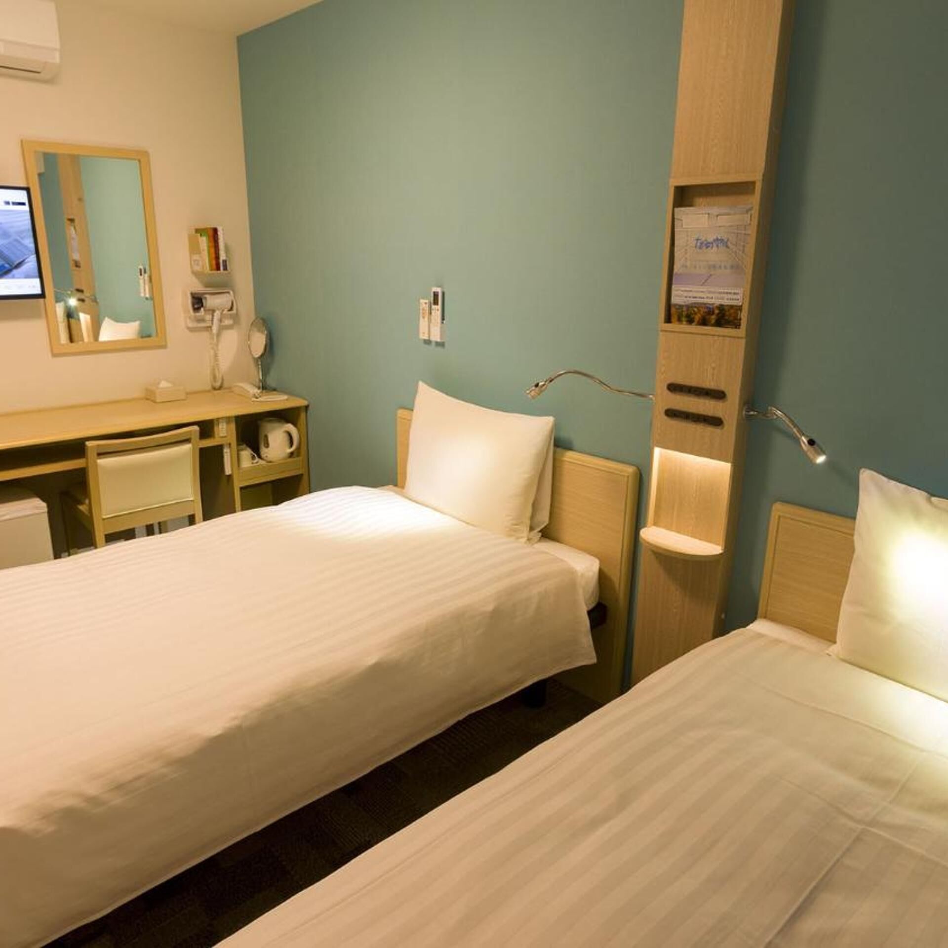 A hotel room with two beds, a desk, a mirror, a phone, and a wall-mounted light fixture.