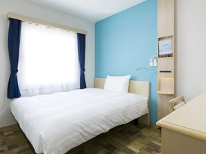 A hotel room with a bed, a desk, a chair, a window with curtains, and a wall-mounted control panel.