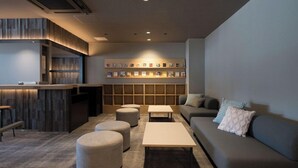 A modern lounge area with a sofa, ottomans, a coffee table, a reception desk, and a wall-mounted bookshelf. - Book Hotel Kyotokujo (Otsu)
