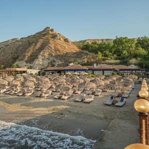 A beach with sun loungers and umbrellas, a mountain in the background, and a building with a tiled roof. - Maria Palace Hotel (Balchik)
