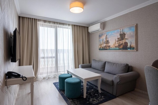 A hotel room with a sofa, a small table, two ottomans, a telephone, a painting of ships, and a window with curtains. - Maria Palace Hotel (Balchik)