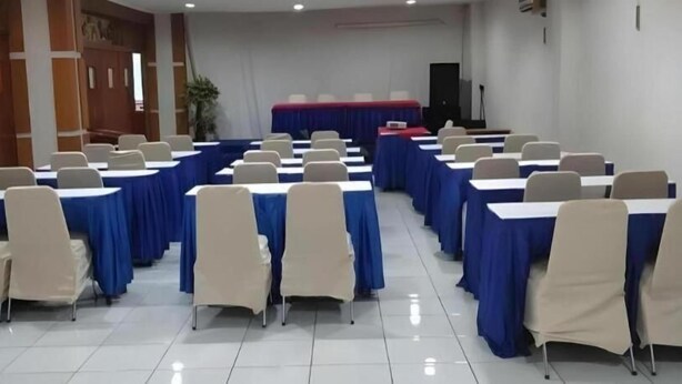 A conference room with rows of tables and chairs, a stage with a red backdrop, and a white tiled floor. - Alexander Hotel Tegal (Tegal)