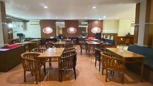 A spacious dining area with multiple wooden tables and chairs, a central bar area, and a seating section with a blue couch. - Alexander Hotel Tegal (Tegal)