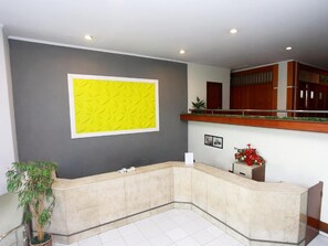 A reception area with a curved reception desk, a large yellow decorative wall, a potted plant, and a wooden counter with a red flower arrangement. - Alexander Hotel Tegal (Tegal)