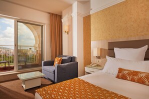 Une chambre d’hôtel avec un grand lit, un canapé, une table basse en verre, une table de chevet et une vue sur la ville par une grande fenêtre.